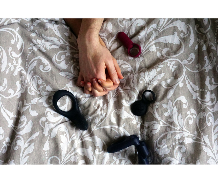 Deux mains entrelacées reposent sur un lit aux motifs élégants, entourées de plusieurs accessoires en silicone. L’image évoque complicité et découverte sensorielle.